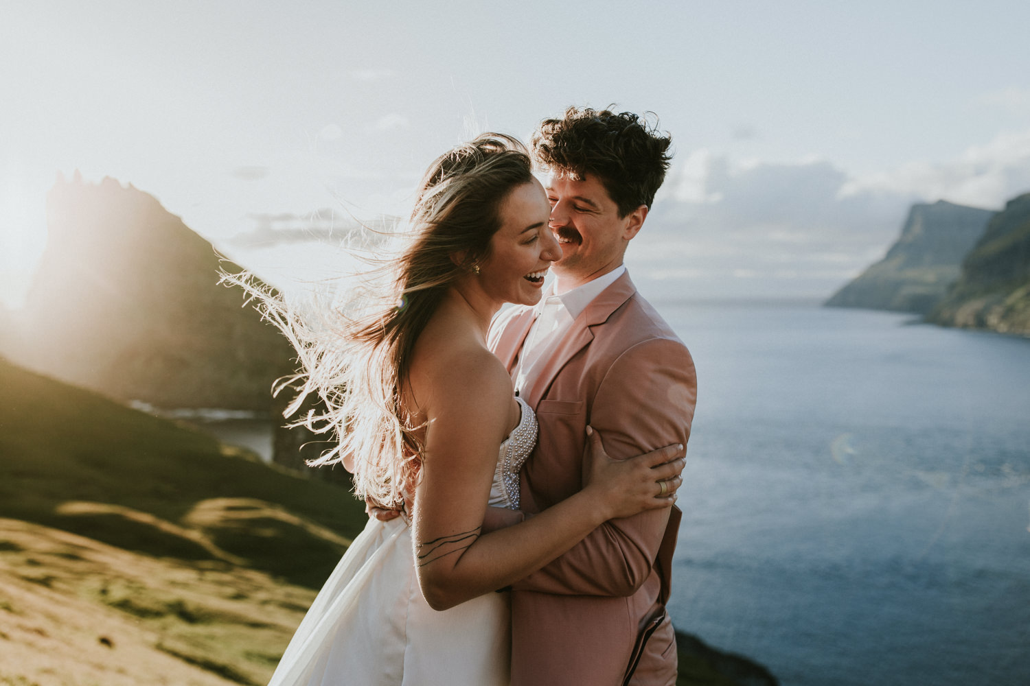 A hetero couple in white wedding dress and pink suit embrace and laugh in front of a dramatic, evening sun and the jagged cliffs of the Faroe Islands behind them.