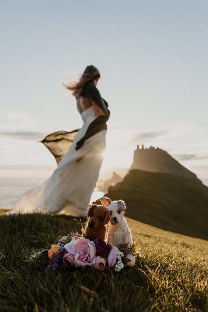 A bride in white wedding dress stands in front of two crochet dogs, facing away from the camera toward a dramatic, golden sunset with the jagged peaks of the Faroe Islands in the distance