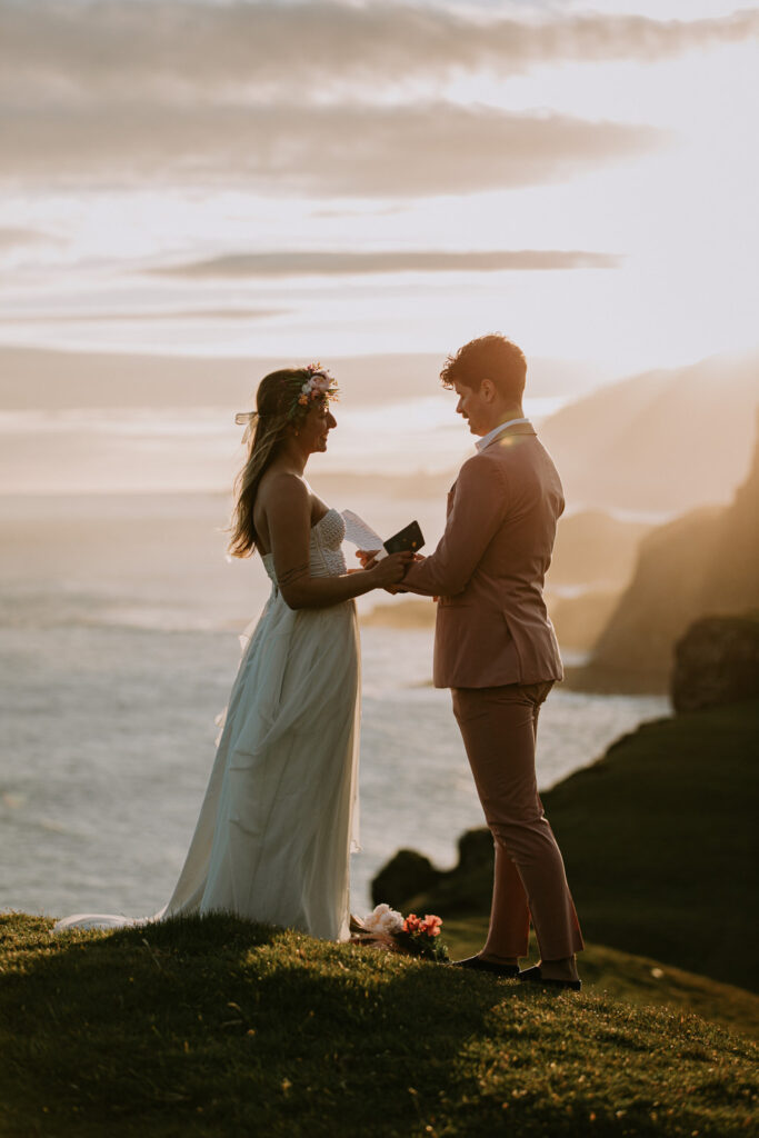 A hetero couple in white wedding dress and pink suit read elopement vows against a dramatic, golden sunset with the misty shores of the Faroe Islands behind them.