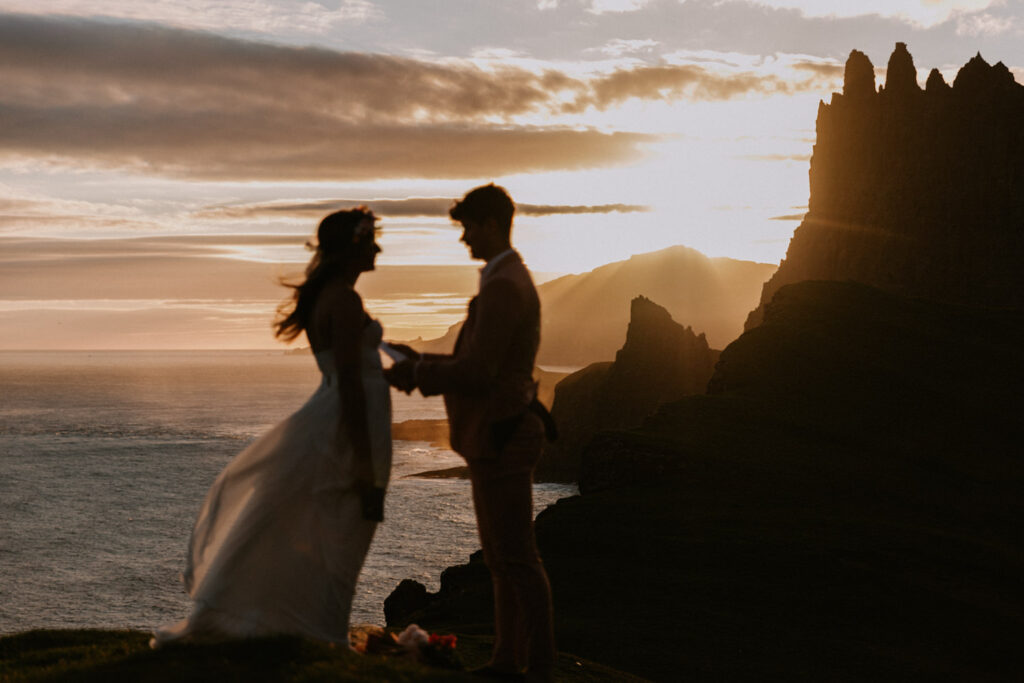 A hetero couple stand in silhouette reading elopement vows against a dramatic, golden sunset with the misty shores of the Faroe Islands behind them.