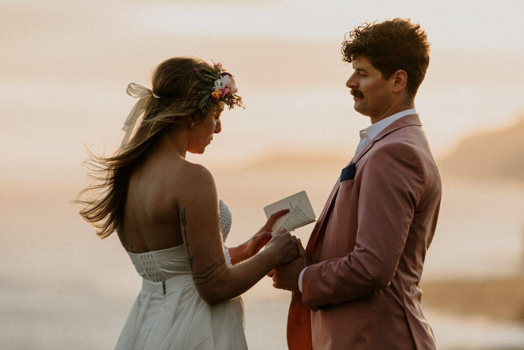 A hetero couple stand reading elopement vows against a dramatic, golden sunset with the misty shores of the Faroe Islands behind them.