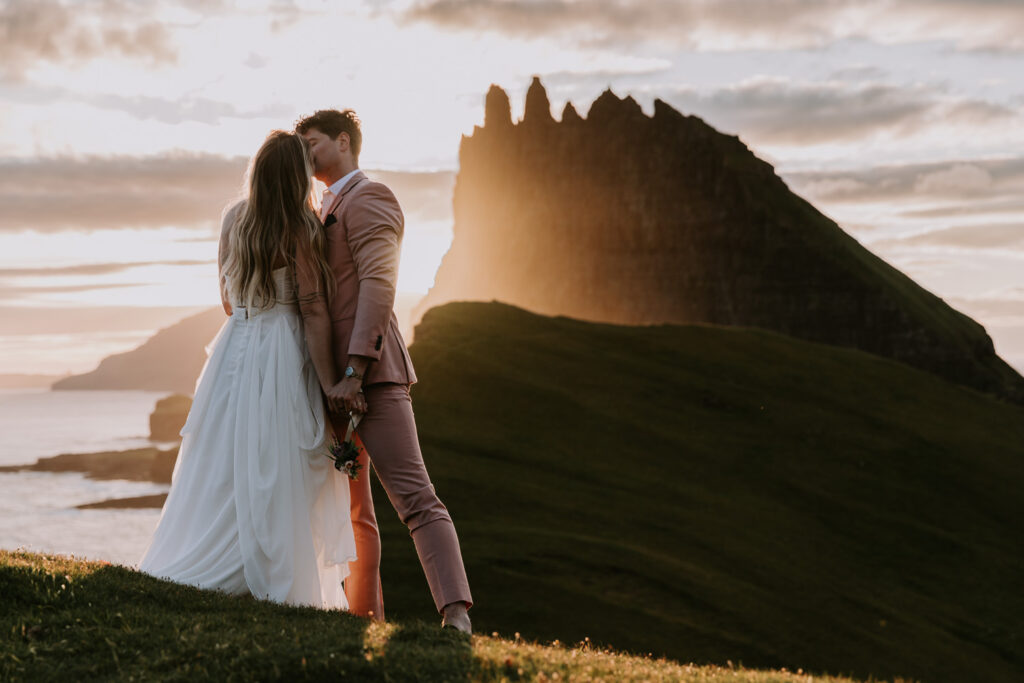 A hetero couple in white wedding dress and pink suit hug against a dramatic, golden sunset with the jagged peaks of the Faroe Islands behind them.