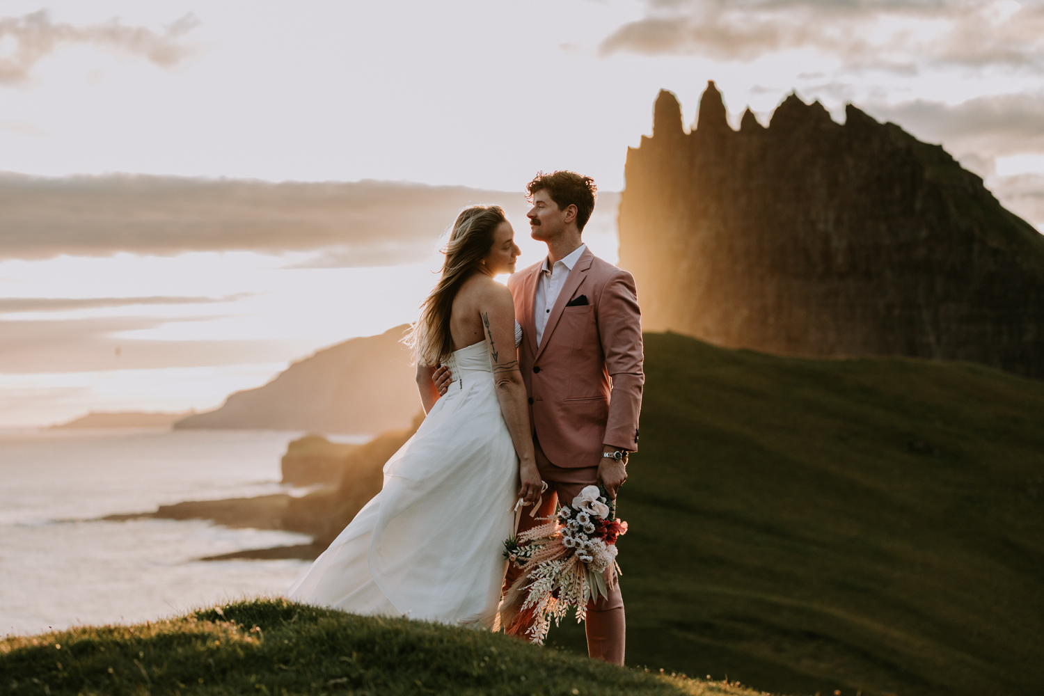 A hetero couple in white wedding dress and pink suit hug against a dramatic, golden sunset with the jagged peaks of the Faroe Islands behind them.