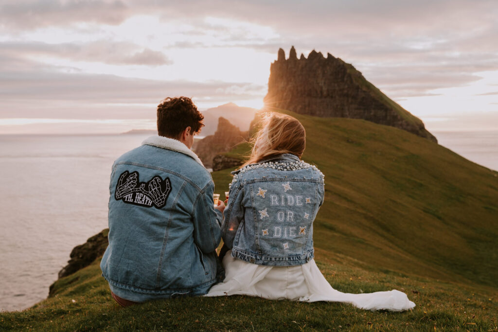 A hetero couple sit in matching jean jackets on their elopement day sipping whiskey and watching a golden sunset with the misty shores of the Faroe Islands in the distance.