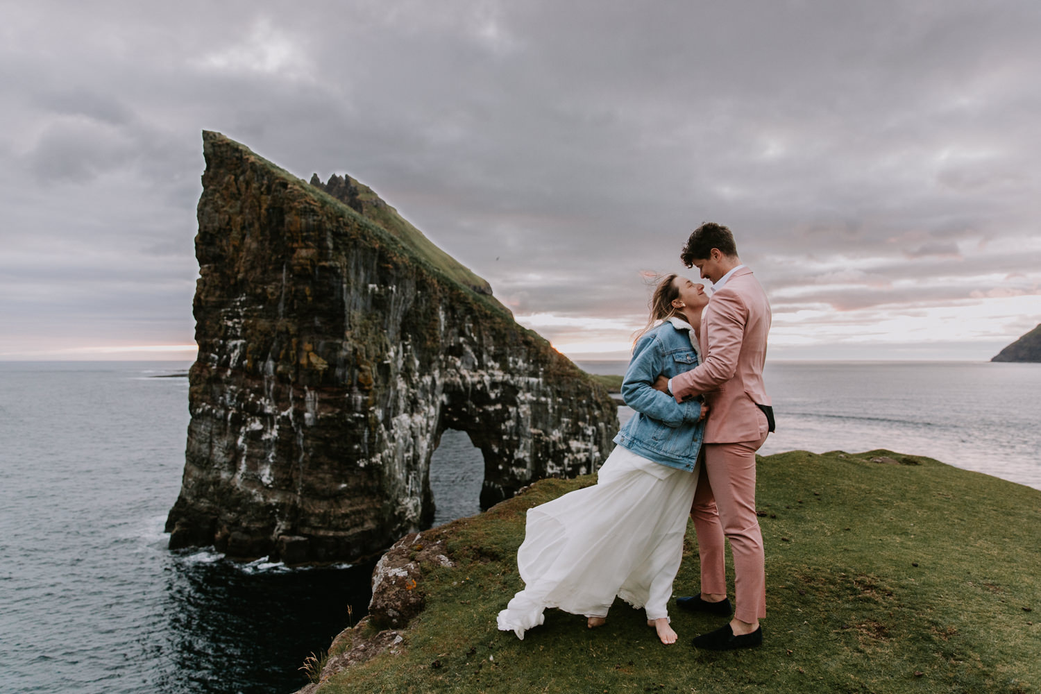 A hetero couple in white wedding dress and jean jacket, and pink suit face each other smiling with the dramatic sea stack of Drangarnir behind them in the Faroe Islands.