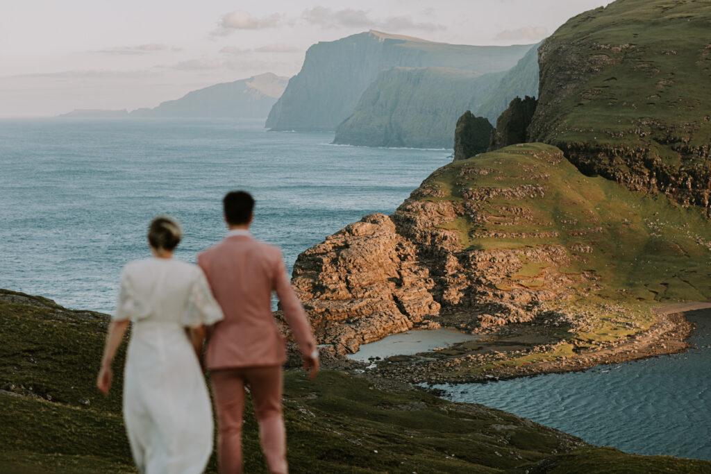 A couple in white wedding dress and pink suit walk away from the camera out of focus against the dramatic, landscape and coastline of the Faroe Islands ahead of them them.