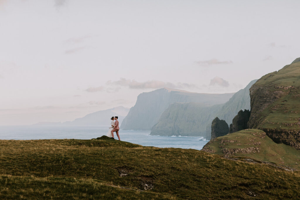 A hetero couple in white wedding dress and pink suit stand far away in the frame against a dramatic, coastal landscape of the Faroe Islands behind them.