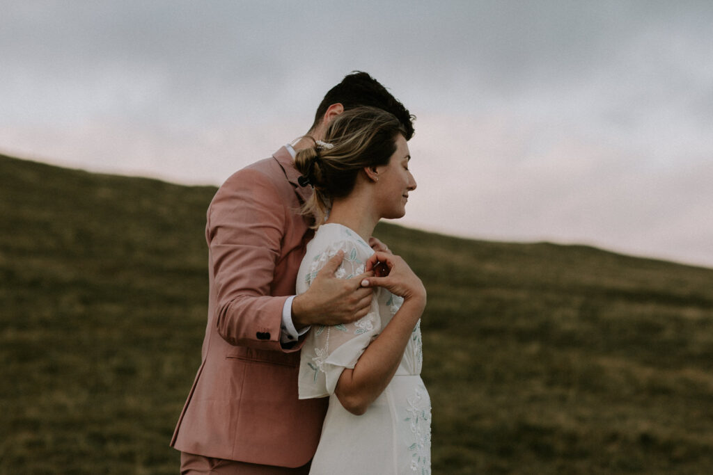A hetero couple in white wedding dress and pink suit stand holding each other against the grassy landscape of the Faroe Islands.