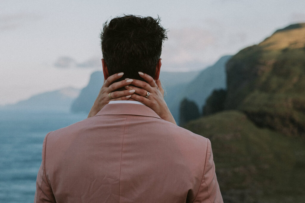 A woman's fingers are seen draped around the back of a her husband's neck with the dramatic, coastal landscape of the Faroe Islands behind them.