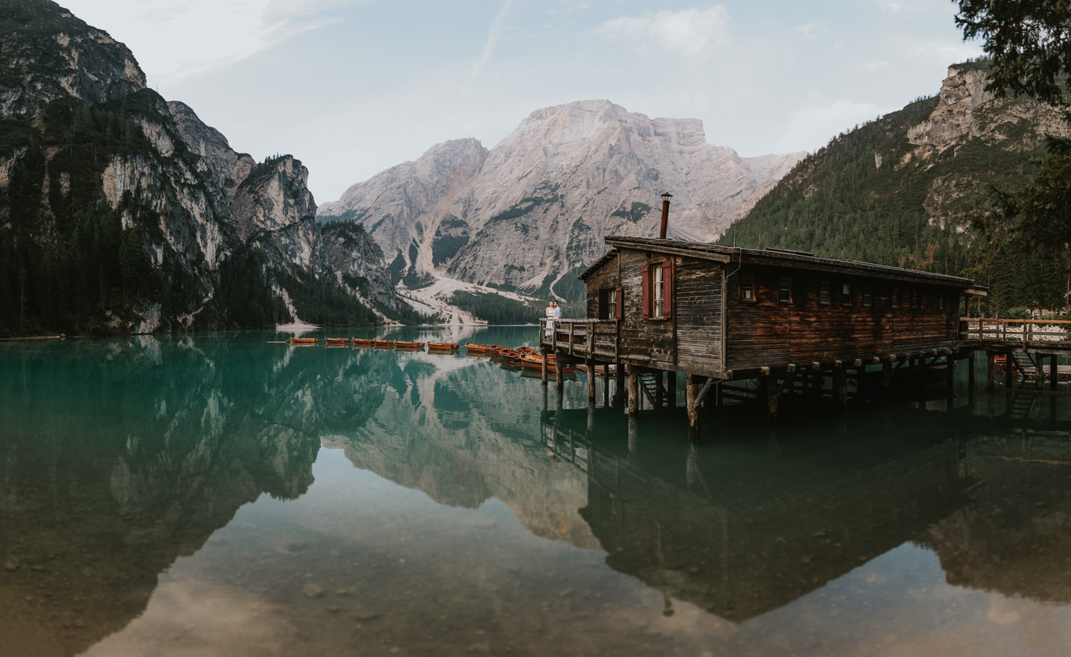 A couple stand far away in the center of the frame. They are standing on the wooden dock of the Lago di Braies boathouse, with the dramatic Dolomites mountains reflected in the crystal clear water.