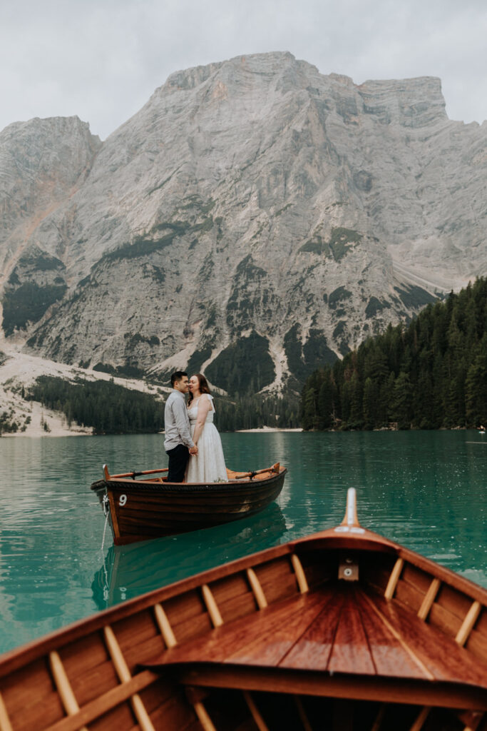 A couple stand on the famous wooden boats on Lago di Braies, one wooden boat in the foreground and the dramatic peaks of the Dolomites in the background.