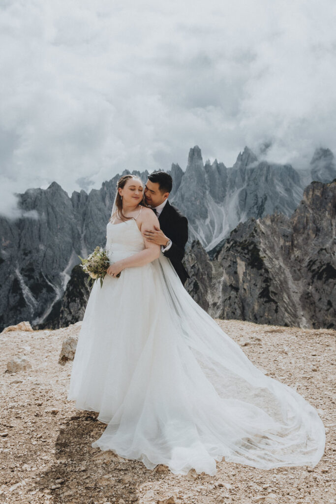 A couple stand in the middle of the frame embracing during their elopement. They are framed by the jagged, mist covered mountain peaks of the Cadini range in the Italian Dolomites. They are wearing a black tuxedo and white wedding dress.