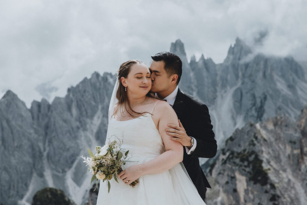 A couple stand kissing in the middle of the frame during their elopement. They are framed by the jagged, mist covered mountain peaks of the Cadini range in the Italian Dolomites. They are wearing a black tuxedo and white wedding dress.