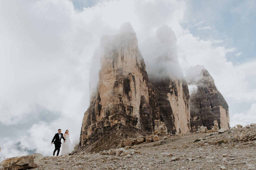A couple in wedding clothes run under the towering Tre Cime mountain peaks, covered in mist and fog on their elopement day.