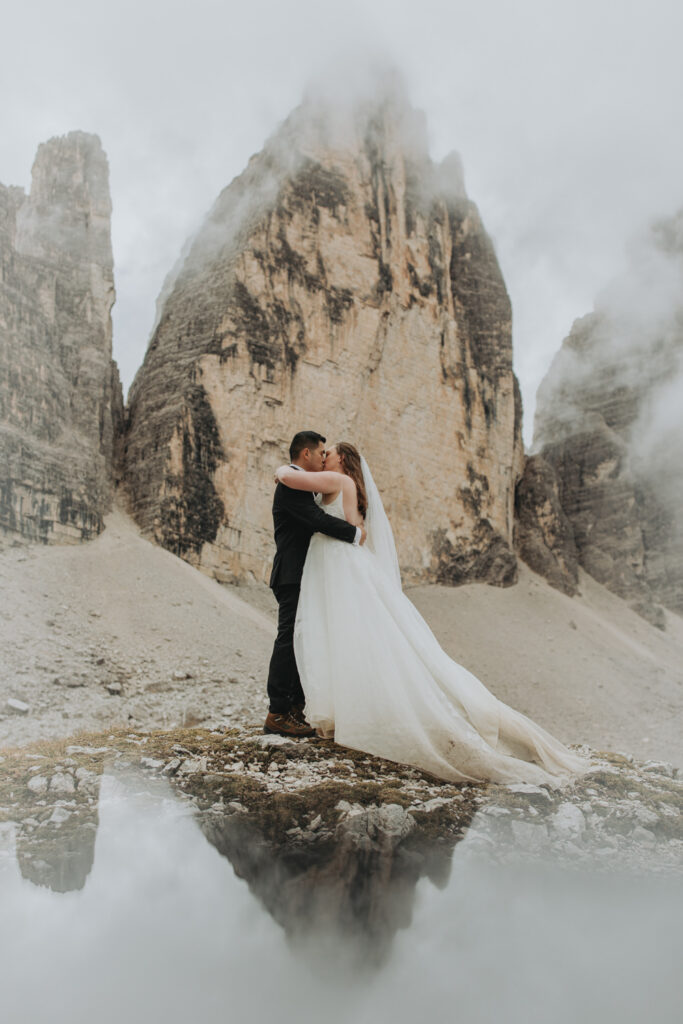 A couple stand in the middle of the frame embracing during their elopement. They are framed by the jagged, mist covered mountain peaks of the Tre Cime massif in the Italian Dolomites. They are wearing a black tuxedo and white wedding dress.