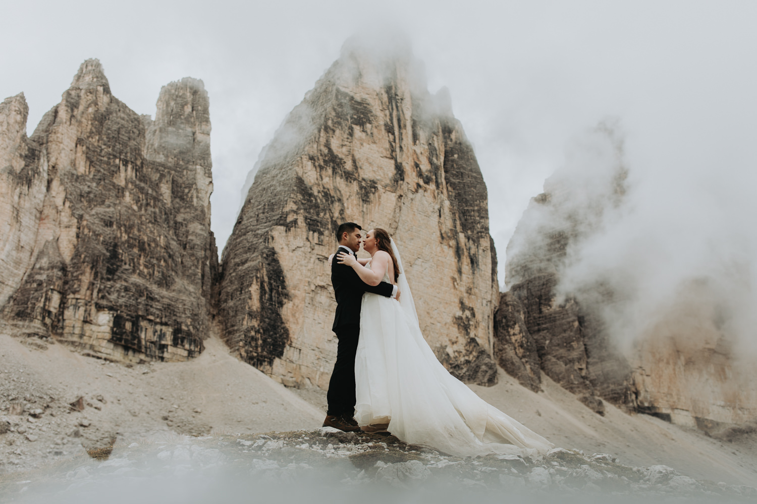 A couple stand in the middle of the frame embracing during their elopement. They are framed by the jagged, mist covered mountain peaks of Tre Cime in the Italian Dolomites. They are wearing a black tuxedo and white wedding dress.