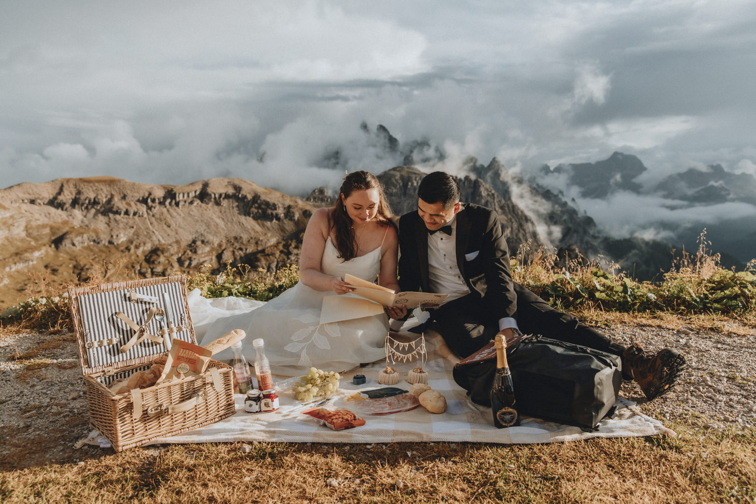 A couple sit having a picnic in the middle of the frame, reading letters during their elopement. They are framed by the jagged, mist covered mountain peaks of the Cadini di Misurina range in Tre Cime national park.