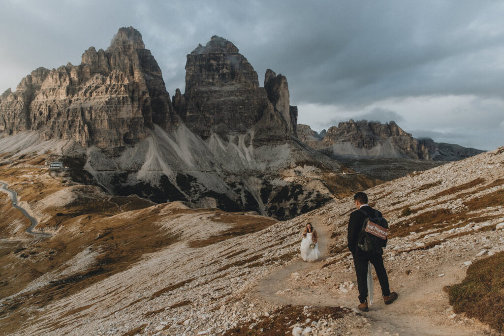 A couple in wedding dress and black tuxedo walk toward the camera on their elopement day in Tre Cime National Park. The famous Tre Cime peaks are behind them, and the sun is setting dramatically to their left.