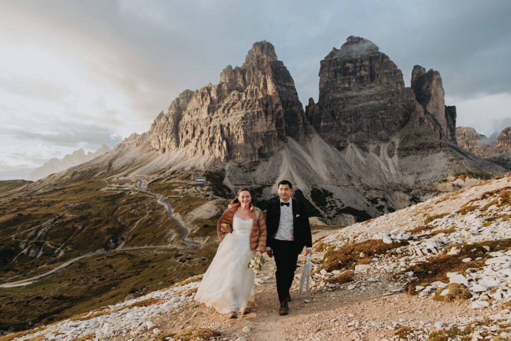A couple in wedding dress and black tuxedo walk toward the camera on their elopement day in Tre Cime National Park, Dolomites. The famous Tre Cime peaks are behind them, and the sun is setting dramatically to their left.