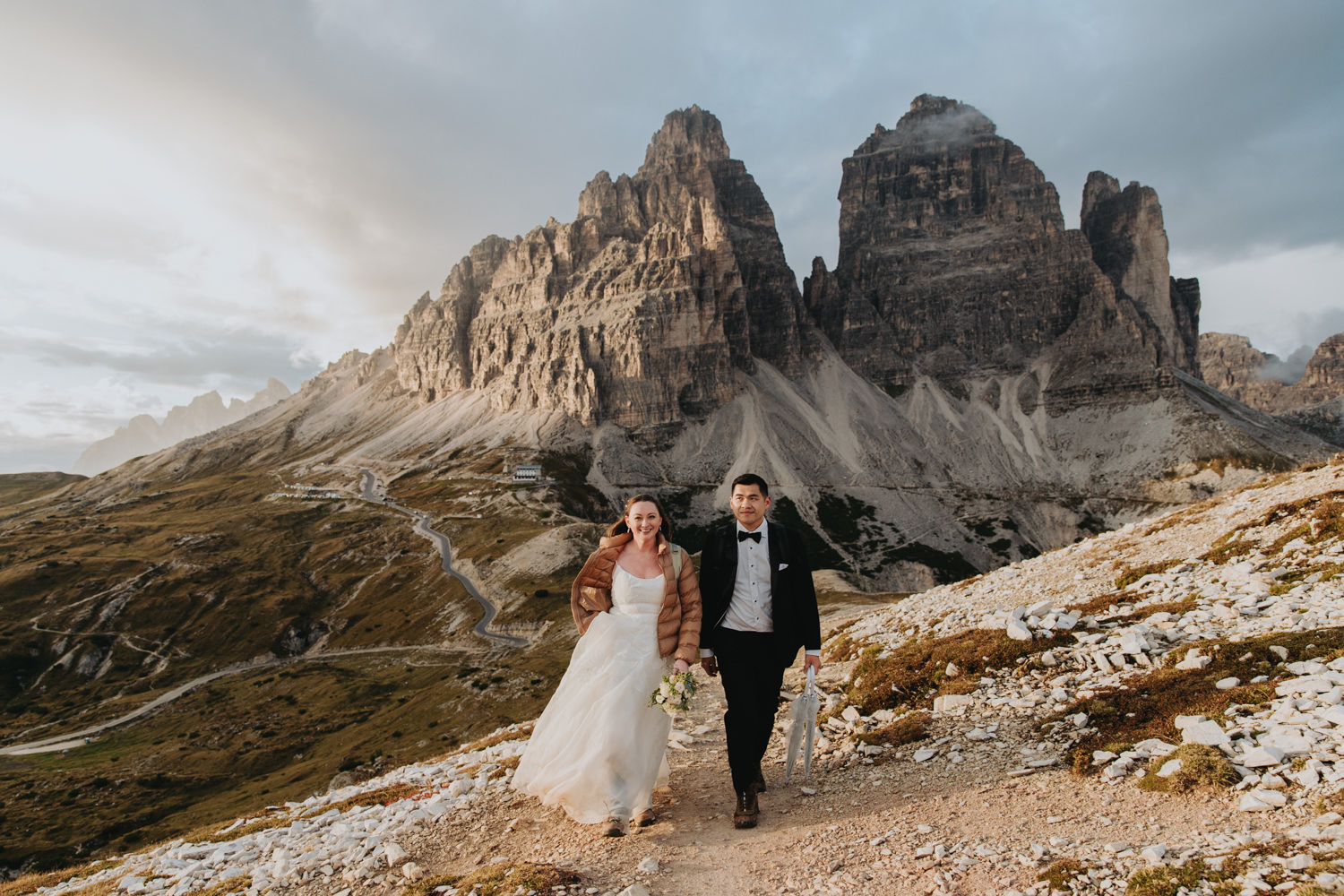 A couple walk in wedding attire at sunset in the Italian Dolomites. The famous Tre Cime is behind them.