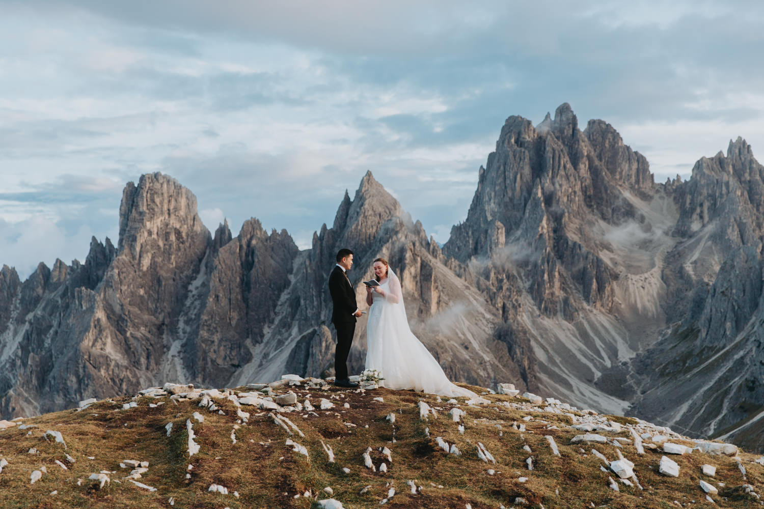 A couple stand in the middle of the frame exchanging wedding vows during their Dolomites elopement. They are framed by the jagged, mist covered mountain peaks of the Cadini di Misurina range in Tre Cime national park.