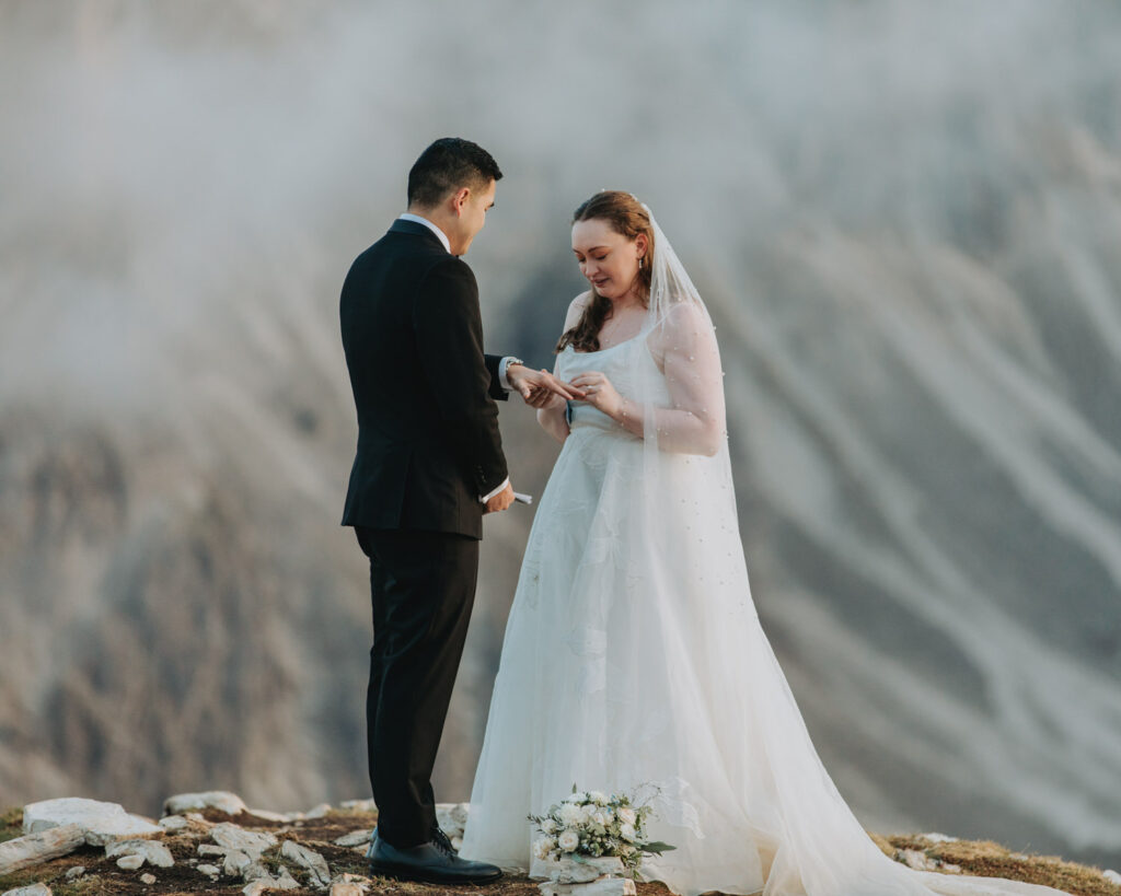A couple stand in the middle of the frame exchanging wedding rings during their elopement. They are framed by the jagged, mist covered mountain peaks of the Cadini di Misurina range in Tre Cime national park.