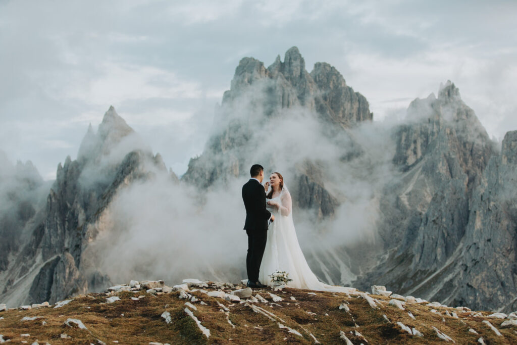 A couple stand in the middle of the frame exchanging wedding vows during their elopement. They are framed by the jagged, mist covered mountain peaks of the Cadini di Misurina range in Tre Cime national park.