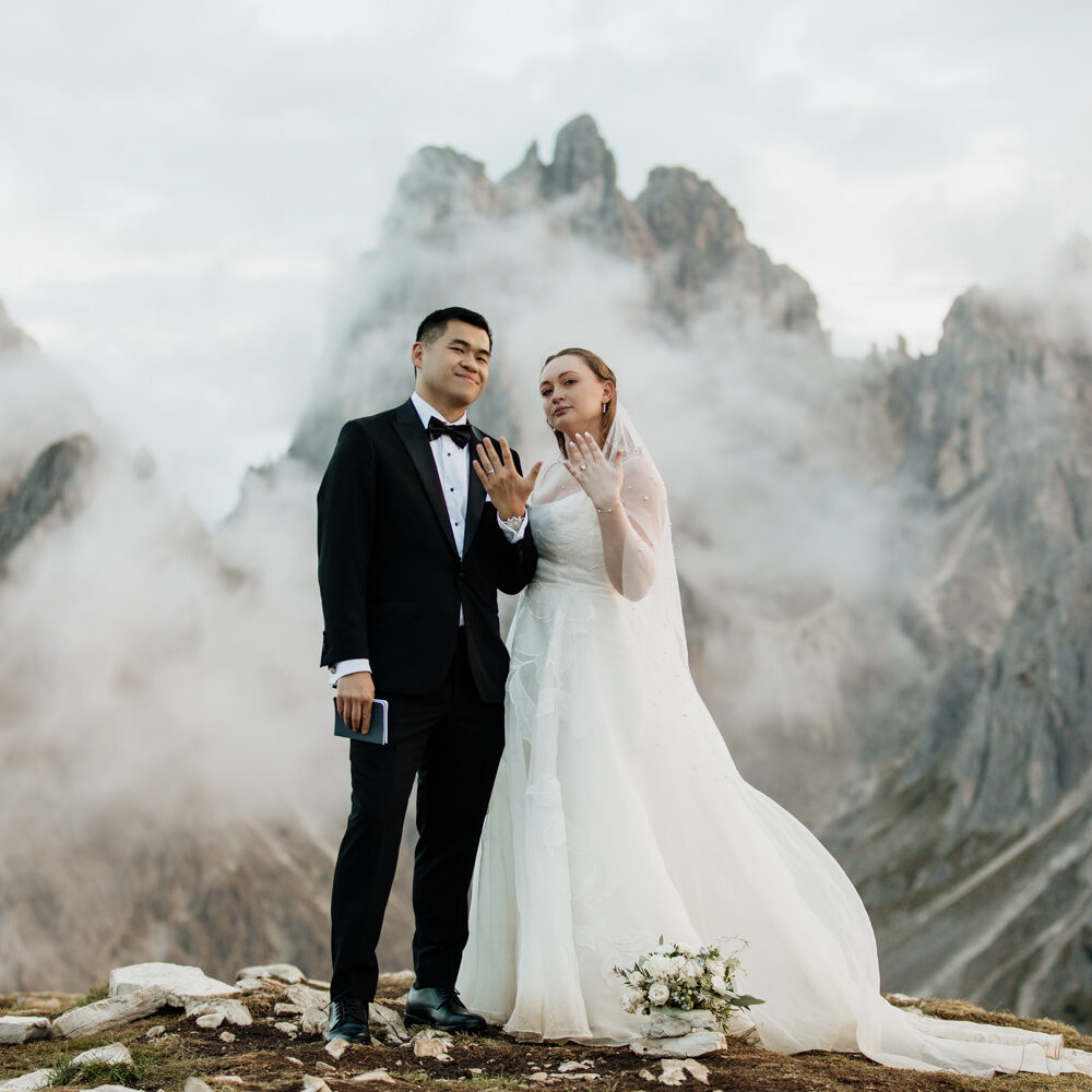 A couple stand in the middle of the frame holding out their hands to show their wedding rings. They are framed by the jagged, mist covered mountain peaks of the Cadini di Misurina range in Tre Cime national park. They are wearing a black tux and wedding dress, posing for their elopement photos.
