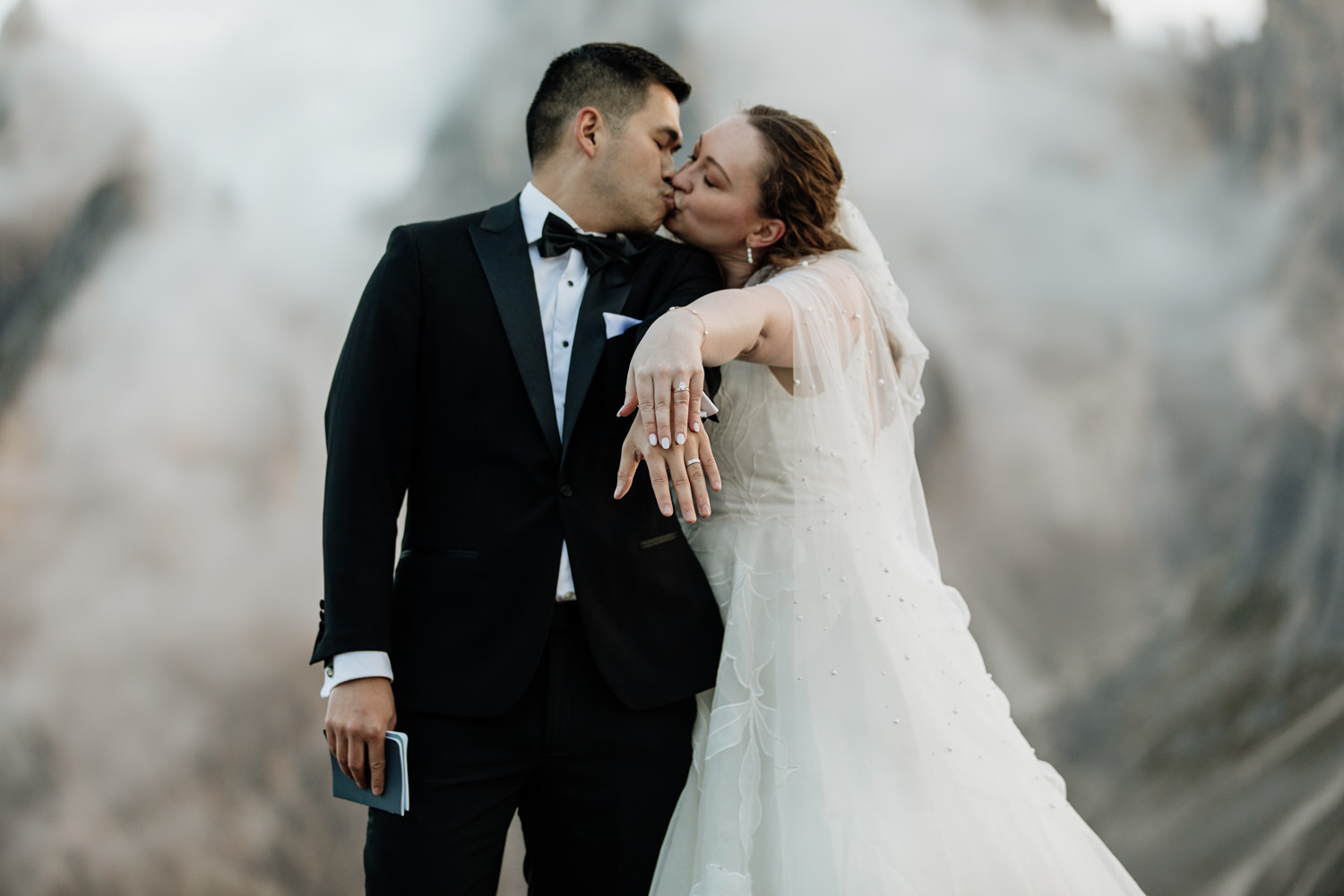 A couple stand in the middle of the frame holding out their hands to show their wedding rings. They are framed by the jagged, mist covered mountain peaks of the Cadini di Misurina range in Tre Cime national park. They are wearing a black tux and wedding dress, posing for their elopement photos.
