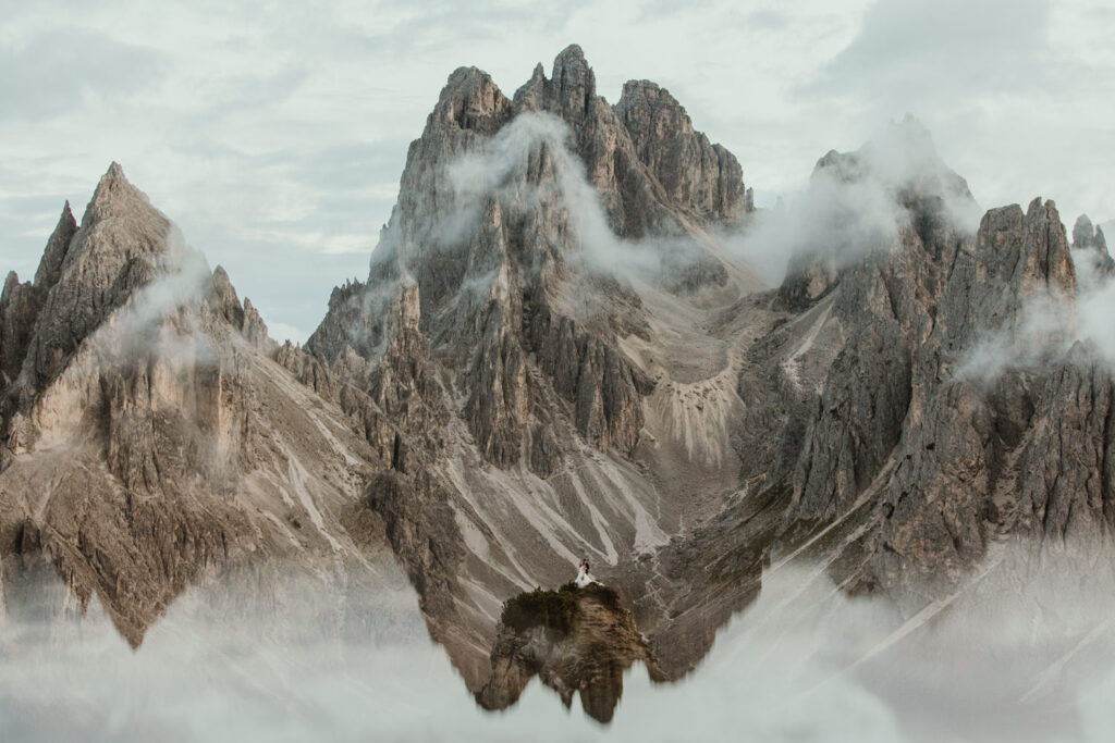 A couple stand far away and tiny in the distance, surrounded by the jagged, mist covered mountain peaks of the Cadini di Misurina range in Tre Cime national park. They are wearing a black tux and wedding dress, posing for their elopement photos.