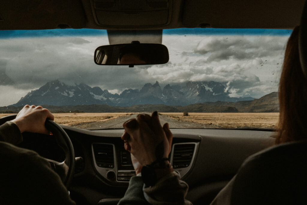 A shot inside a car looking out the windshield toward the peaks of Torres del Paine National Park in Patagonia. A couple is out of focus holding hands in the driver and passenger seat.