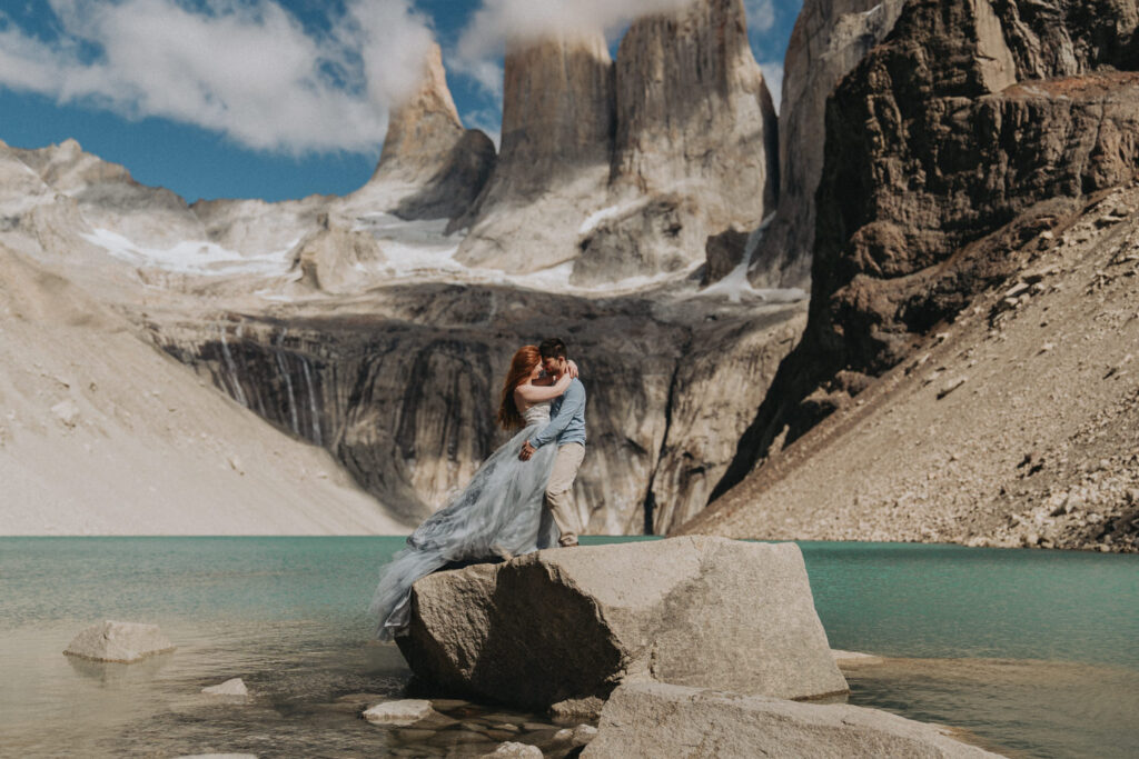 A couple hug on top of a large boulder in Torres del Paine National Park on their elopement day. There are towering rock pillars and a teal blue lake behind them.