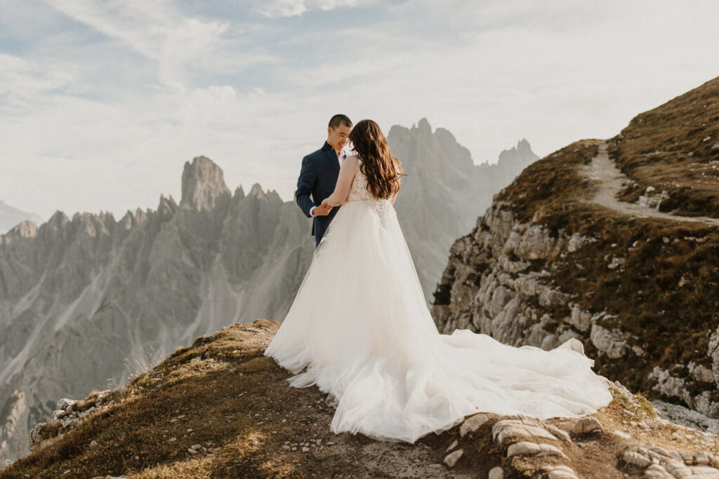 An Asian couple embrace in wedding clothes in front of the dramatic Cadini di Misurina mountain range during a golden autumn evening, posing for their elopement photos. 
