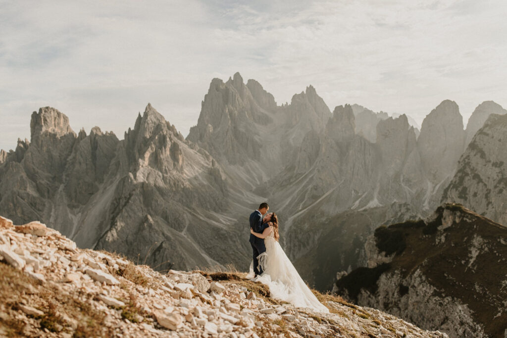 A couple in wedding attire stand on a mountain side in the Italian Dolomites, leaning in for a kiss with the famous Cadini di Misurina peaks behind them at sunset