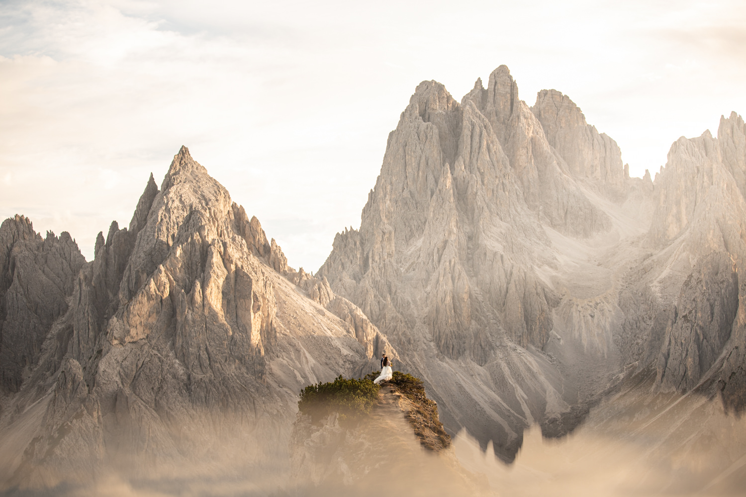 A couple in wedding clothes stand far in the frame under the dramatic Cadini di Misurina mountain range at sunset, posing for their elopement photos. 