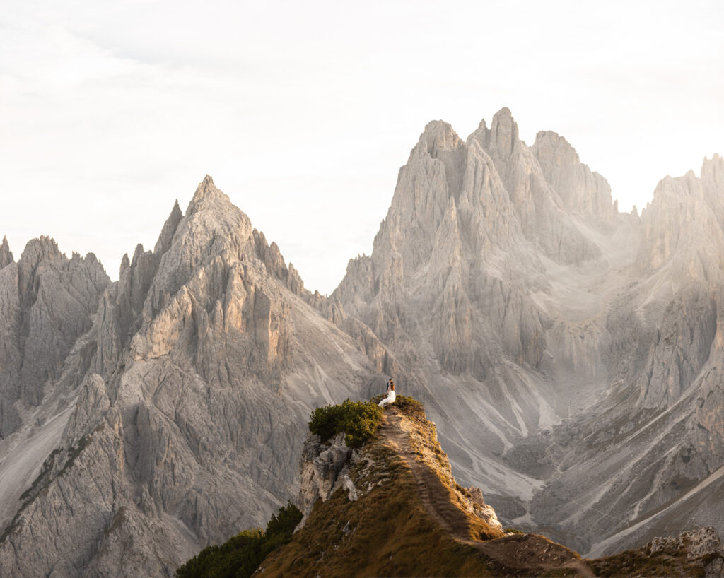 A couple in wedding clothes stand far in the frame under the dramatic Cadini di Misurina mountain range at sunset, posing for their elopement photos. 
