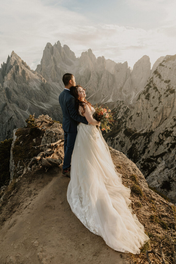 An Asian couple embrace in wedding clothes in front of the dramatic Cadini di Misurina mountain range during a golden sunset, posing for their elopement photos. 