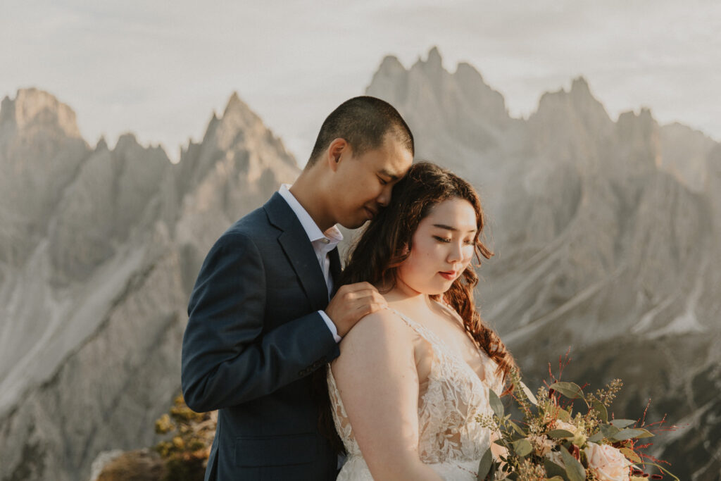 An Asian couple embrace with their eyes closed in wedding clothes in front of the dramatic Cadini di Misurina mountain range during a golden sunset, posing for their elopement photos. 