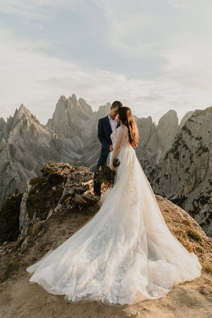 An Asian couple embrace in wedding clothes in front of the dramatic Cadini di Misurina mountain range during a golden sunset, posing for their elopement photos. 