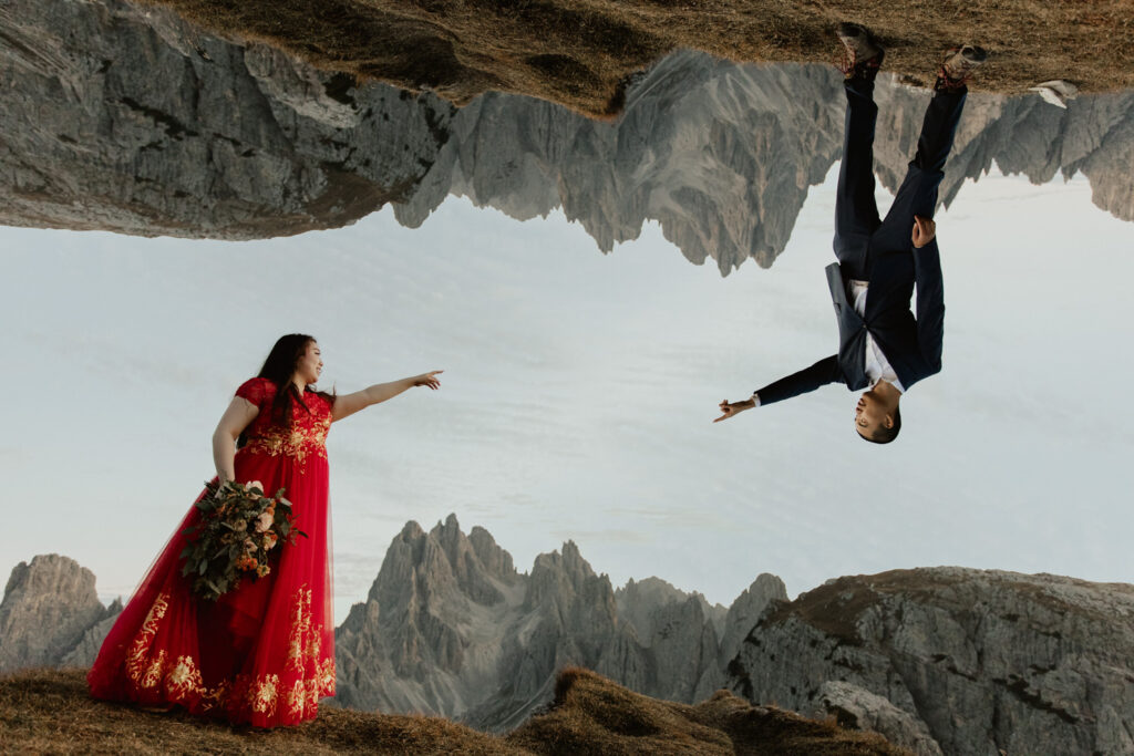 An Asian couple pose for a trippy inverted mountain range image in a traditional red Chinese dress and blue suit. They are pointing at each other in front of the dramatic Cadini di Misurina mountain range for their elopement photos. 