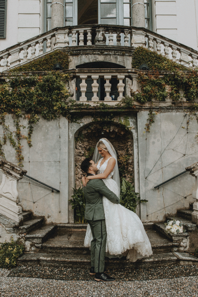 A groom picks up his wife at Villa Carlotta for their lake Como elopement. They are framed by red flowers, fountains, and draping greenery in the villa gardens.