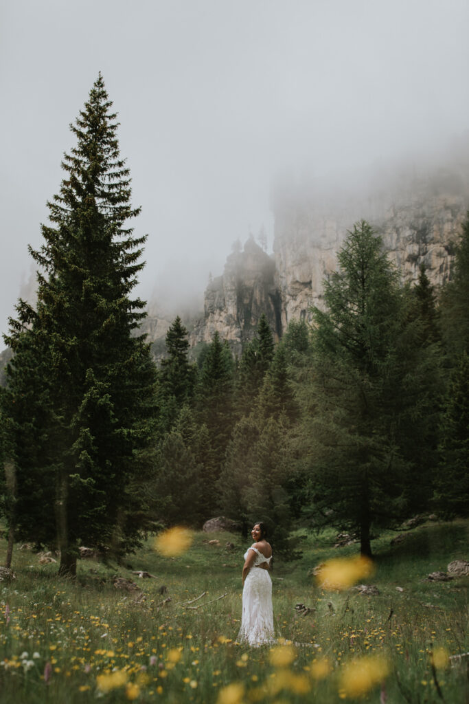 A woman in a white dress poses for the camera in a field of wildflowers on a misty July day in the Italian Dolomites