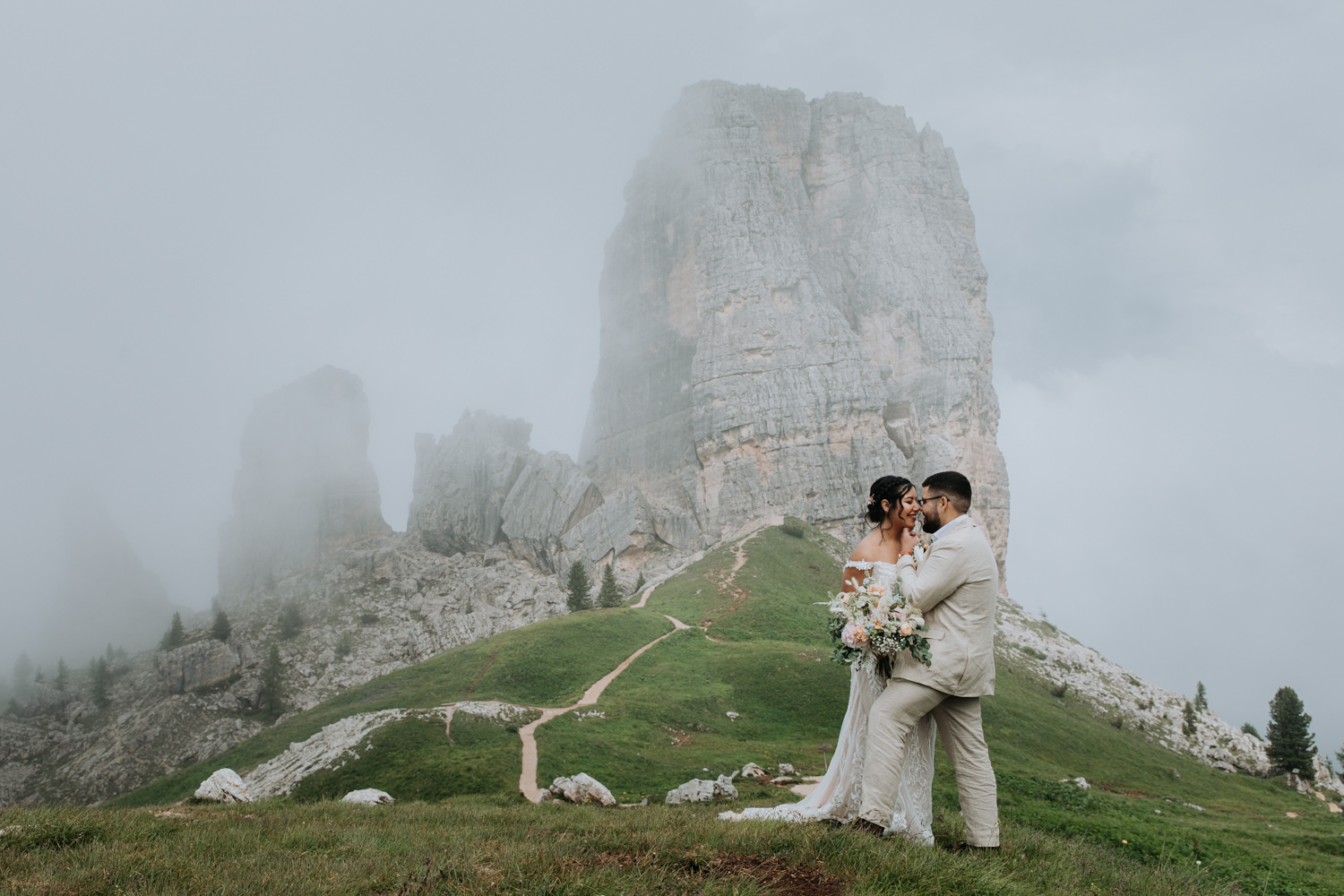A couple in champagne colored wedding dress and linen suit stand on top of a grassy and misty mountain in the Dolomites during their July elopement, nuzzling noses.