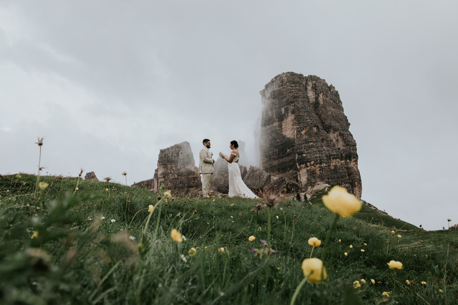 A couple stand in front of the rocky spire of Cinque Torri in the Dolomites, reading elopement vows. They are framed by big yellow flowers in the foreground.