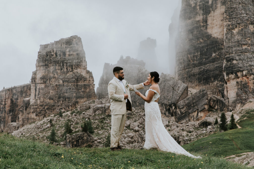A couple in champagne colored wedding dress and linen suit read vows on top of a misty, grassy mountain in the Dolomites during their July elopement