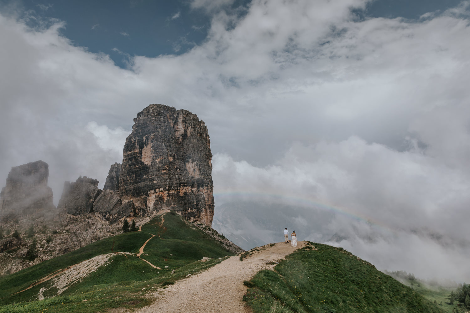 A couple in champagne colored wedding dress and linen suit walk down a gravel trail toward a tall rocky mountain peak and a rainbow in the Dolomites during their July elopement