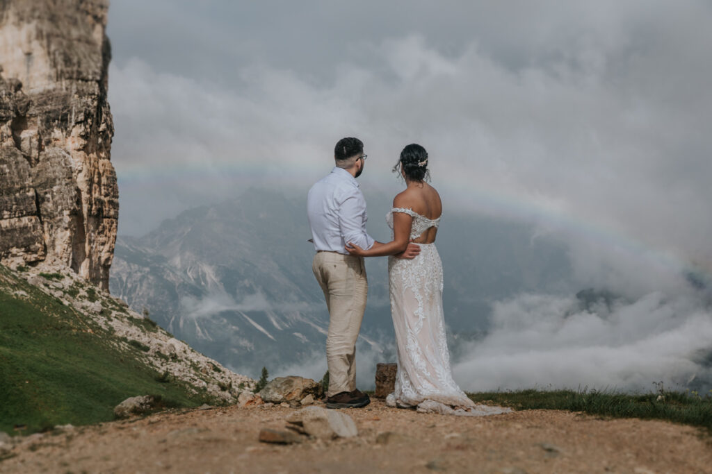 A couple in white wedding attire stares at a rainby next to a rocky spire in the Dolomites during their July elopement