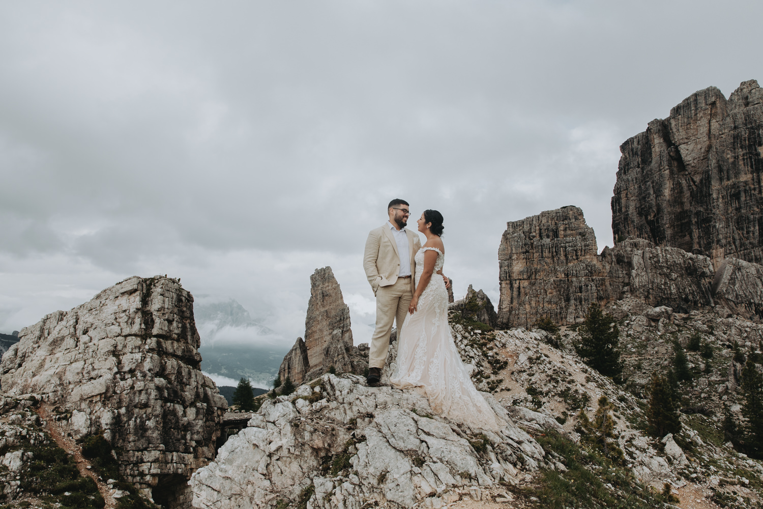 A couple in champagne colored wedding dress and linen suit stand on top of a rocky outcropping in the Dolomites during their July elopement, looking at one another