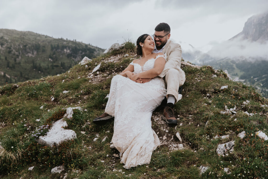 A couple in champagne colored wedding dress and linen suit sit on top of a grassy mountain in the Dolomites during their July elopement, cuddling and smiling at one another