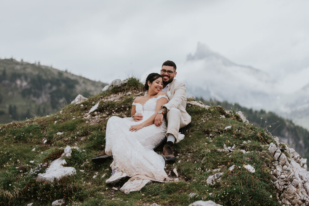 A couple in champagne colored wedding dress and linen suit sit on top of a grassy mountain in the Dolomites during their July elopement, cuddling and smiling
